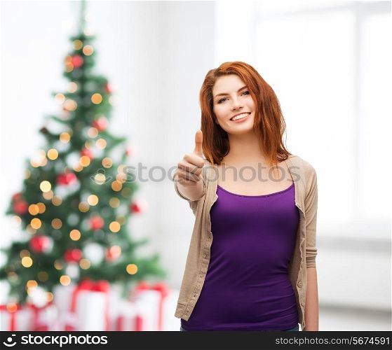 winter holidays, gesture and people concept - smiling teenage girl in casual clothes showing thumbs up over living room and christmas tree background