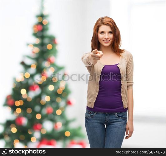 winter holidays, gesture and people concept - smiling teenage girl in casual clothes pointing finger on you over living room and christmas tree background