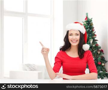 winter, holidays, happiness and people concept - smiling woman in santa helper hat pointing finger over living room with christmas tree background