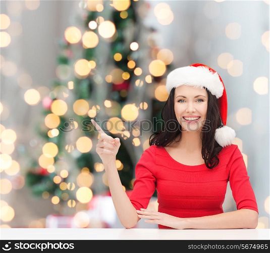 winter, holidays, happiness and people concept - smiling woman in santa helper hat pointing finger over christmas tree lights background