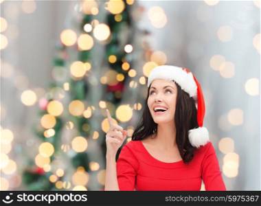 winter, holidays, happiness and people concept - smiling woman in santa helper hat pointing finger over christmas tree lights background