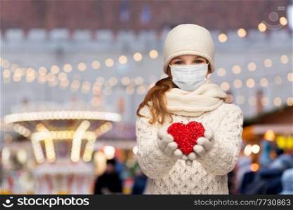 winter holidays, health and charity concept - smiling girl in mask with red heart decoration over christmas market or amusement park lights on background. girl in mask with heart over christmas market