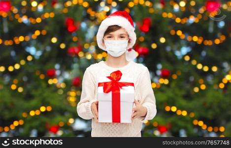winter holidays, health and pandemic concept - boy with gift box wearing face protective medical mask for protection from virus disease over christmas tree lights background. boy in protective mask with christmas gift