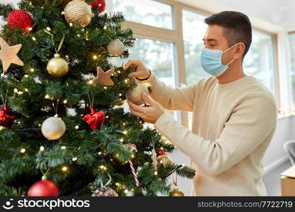 winter holidays, health and people concept - middle aged man in medical mask decorating christmas tree at home. man in mask decorating christmas tree at home