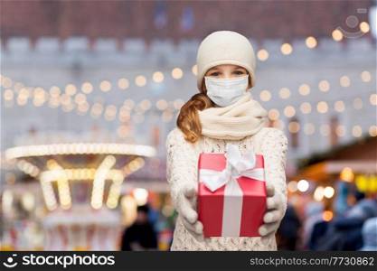 winter holidays, health and people concept - smiling girl in mask with gift box over christmas market or amusement park lights on background. girl in mask with gift box over christmas market