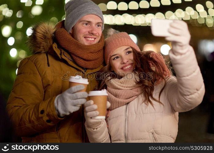 winter holidays, hot drinks and people concept - happy couple with coffee taking selfie in christmas evening. couple with coffee taking selfie at christmas