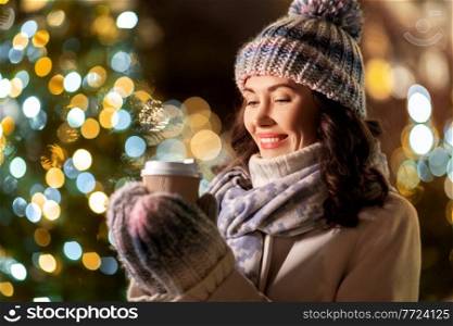 winter holidays, hot drinks and people concept - happy smiling young woman drinking takeaway coffee over christmas tree lights outdoors. happy woman drinking coffee over christmas lights