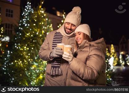winter holidays, hot drinks and people concept - happy young couple with takeaway coffee cups over christmas tree lights in evening city. happy young couple with coffee in christmas city