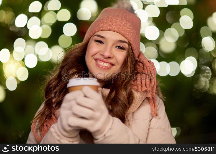 winter holidays, hot drinks and people concept - happy young woman with coffee over christmas tree lights outdoors. happy woman with coffee over christmas lights