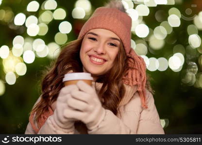 winter holidays, hot drinks and people concept - happy young woman with coffee over christmas tree lights outdoors. happy woman with coffee over christmas lights