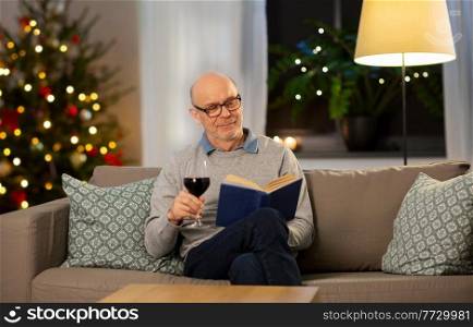 winter holidays, leisure and people concept - happy senior man with glass of red wine reading book at home in evening over christmas tree lights on background. senior man with book drinking wine on christmas