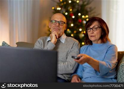 winter holidays, leisure and people concept - senior couple with remote control watching tv at home in evening over christmas tree lights on background. senior couple watching tv at home on christmas