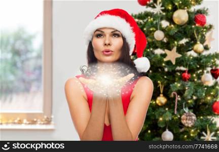 winter holidays, magic and celebration concept - beautiful woman in santa hat and red dress blowing fairy dust off over christmas tree at home on background. woman blowing off fairy dust over christmas tree