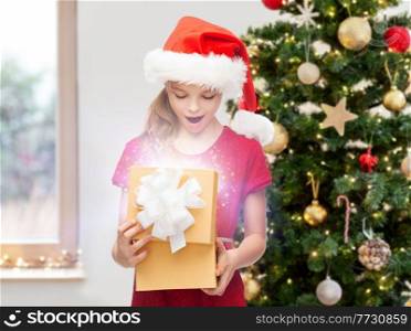 winter holidays, magic and children concept - smiling girl in santa helper hat opening gift box with fairy dust at home over christmas tree background. smiling girl in santa hat with christmas gift