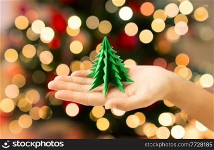 winter holidays, new year and celebration concept - close up of hand holding green paper origami christmas tree over bokeh lights on background. hand holding green paper origami christmas tree