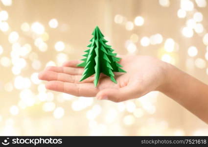 winter holidays, new year and craft concept - hand holding green paper origami christmas tree over festive lights background. hand holding green paper origami christmas tree