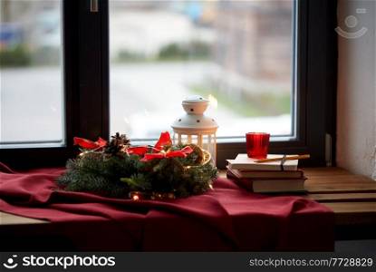 winter holidays, new year and decorations concept - christmas fir wreath, books, candle and lantern on window sill at home. christmas wreath, books, candle, lantern on window