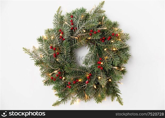 winter holidays, new year and decorations concept - wreath of fir branches with red berries and garland lights on white background. christmas fir wreath with berries and lights