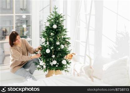 Winter holidays, New Year and people concept. Brunette woman in oversized sweater and jeans decorates Christmas tree, her pedigree dog poses near in spacious white bedroom. Domestic atmosphere