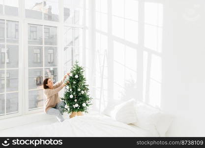 Winter holidays, New Year concept. Pleased young brunette woman has festive mood, decorates Christmas tree at home, prepares for celebration, poses in cozy bedroom with white walls on windowsill.