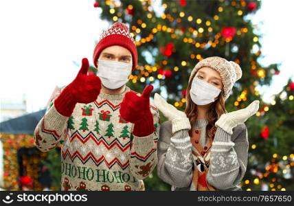 winter holidays, pandemic and health concept - couple in sweaters wearing face protective medical masks for protection from virus disease over christmas tree lights on background. couple in protective masks and christmas sweaters