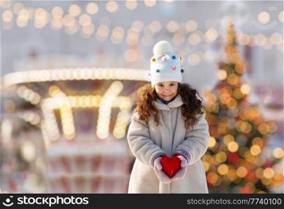 winter holidays, people and celebration concept - happy little girl holding toy heart over christmas tree lights and amusement park background. happy little girl with heart over christmas lights