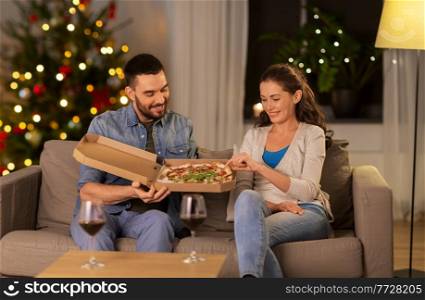 winter holidays, people and fast food concept - happy couple eating takeaway pizza at home in evening over christmas tree lights background. happy couple eating takeaway pizza on christmas