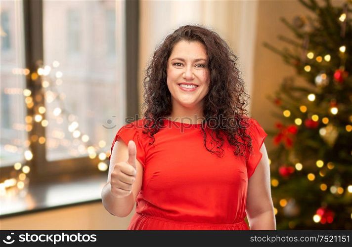 winter holidays, people and gesture concept - happy woman in red dress showing thumbs up over christmas tree lights on home background. happy woman showing thumbs up over christmas tree
