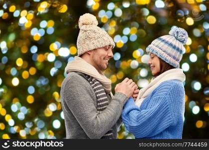 winter holidays, people and love concept - happy romantic couple in knitted hats and scarves holding hands over christmas lights background. happy couple in winter clothes holding hands