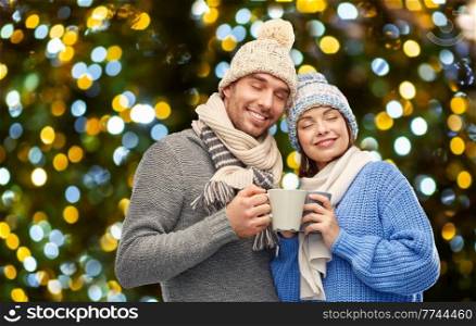 winter holidays, people and love concept - happy romantic couple in knitted hats and scarves with mugs over christmas lights background. happy couple in winter clothes with mugs