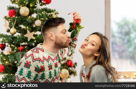 winter holidays, people and traditions concept - happy couple in ugly sweaters kissing under the mistletoe over christmas tree on background. couple kissing under the mistletoe on christmas