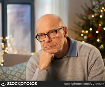 winter holidays, problems and people concept - sad senior man in glasses thinking at home in evening over christmas tree lights on background. sad senior man in glasses at home on christmas