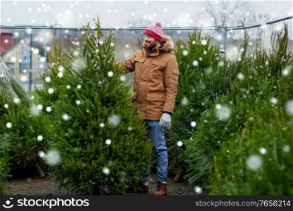 winter holidays, sale and people concept - happy smiling man choosing or selling christmas tree at street market over snow. happy man choosing christmas tree at market