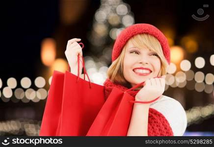 winter holidays, sale and people concept - smiling young woman in hat and scarf with red shopping bags over christmas tree lights background. happy woman with shopping bags over christmas tree