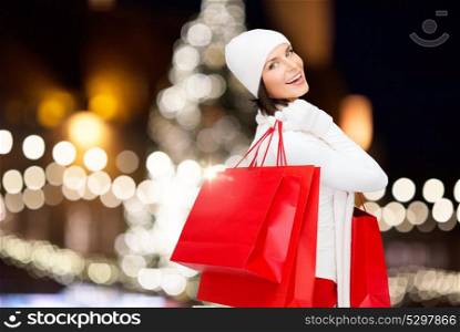 winter holidays, sale and people concept - smiling young woman in white hat and mittens with red shopping bags over christmas tree lights background. happy woman with shopping bags over christmas tree