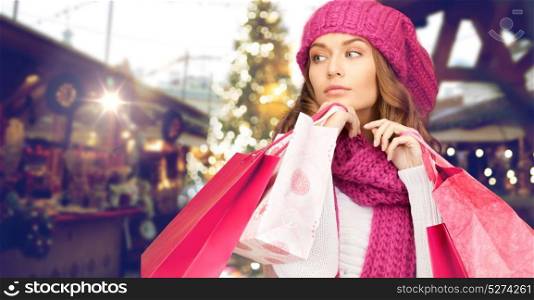 winter holidays, sale and people concept - young woman in hat and scarf with shopping bags over christmas market lights background. happy woman with shopping bags over christmas fair