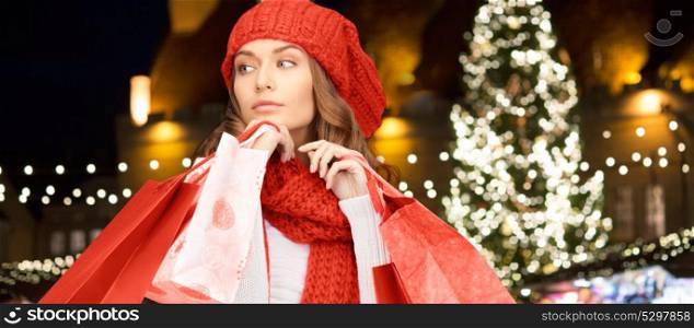 winter holidays, sale and people concept - young woman in hat and scarf with red shopping bags over christmas tree lights background. happy woman with shopping bags over christmas tree