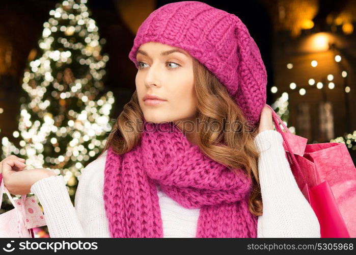 winter holidays, sale and people concept - young woman in hat and scarf with shopping bags over christmas tree lights background. happy woman with shopping bags over christmas tree