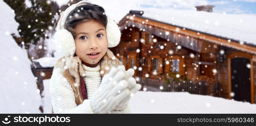 winter holidays, season, christmas, people and children concept - happy little girl wearing earmuffs and gloves over wooden country house background and snow