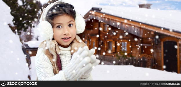 winter holidays, season, christmas, people and children concept - happy little girl wearing earmuffs and gloves over wooden country house background and snow