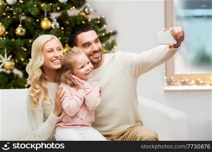 winter holidays, technology and family ople concept - happy father, mother and little daughter sitting on sofa and taking selfie picture with smartphone at home over christmas tree background. family taking selfie with smartphone at christmas