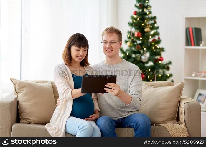 winter holidays, technology and people concept - happy husband and his pregnant wife with tablet pc computer home at christmas. man and pregnant wife with tablet pc at christmas