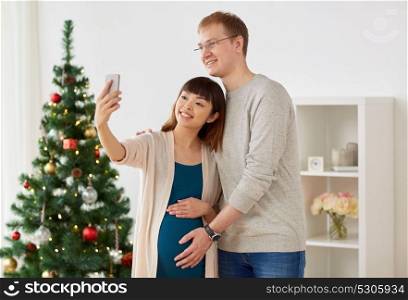 winter holidays, technology and people concept - happy husband and his pregnant wife taking selfie by smartphone home at christmas. happy family couple taking selfie at christmas