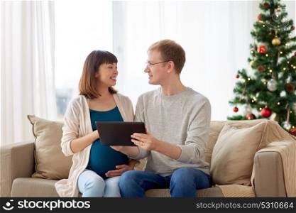 winter holidays, technology and people concept - happy husband and his pregnant wife with tablet pc computer home at christmas. man and pregnant wife with tablet pc at christmas