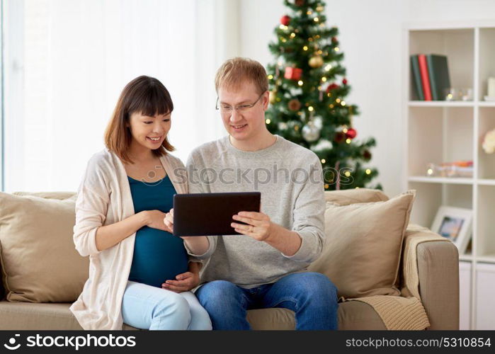 winter holidays, technology and people concept - happy husband and his pregnant wife with tablet pc computer home at christmas. man and pregnant wife with tablet pc at christmas