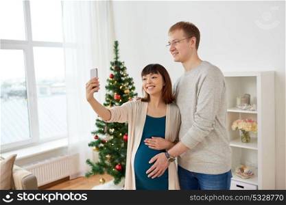 winter holidays, technology and people concept - happy husband and his pregnant wife taking selfie by smartphone home at christmas. happy family couple taking selfie at christmas