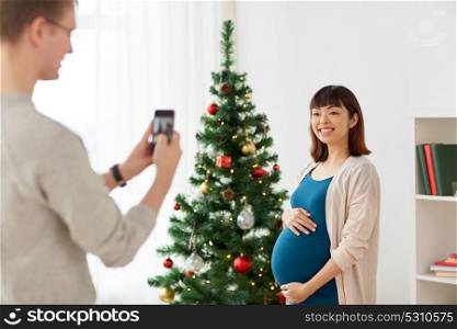 winter holidays, technology and people concept - happy husband photographing his pregnant wife by smartphone at home at christmas. husband photographing pregnant fife at christmas