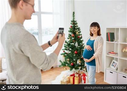 winter holidays, technology and people concept - happy husband photographing his pregnant wife by smartphone at home at christmas. husband photographing pregnant fife at christmas