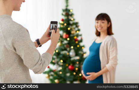 winter holidays, technology and people concept - happy husband photographing his pregnant wife by smartphone at home at christmas. husband photographing pregnant fife at christmas