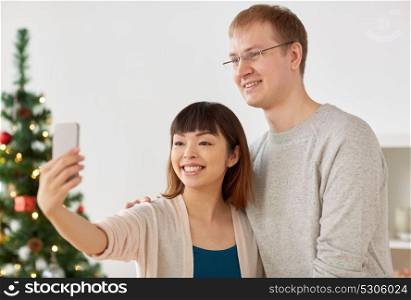 winter holidays, technology and people concept - happy mixed-raced couple taking selfie by smartphone home at christmas. mixed-raced couple taking selfie at christmas